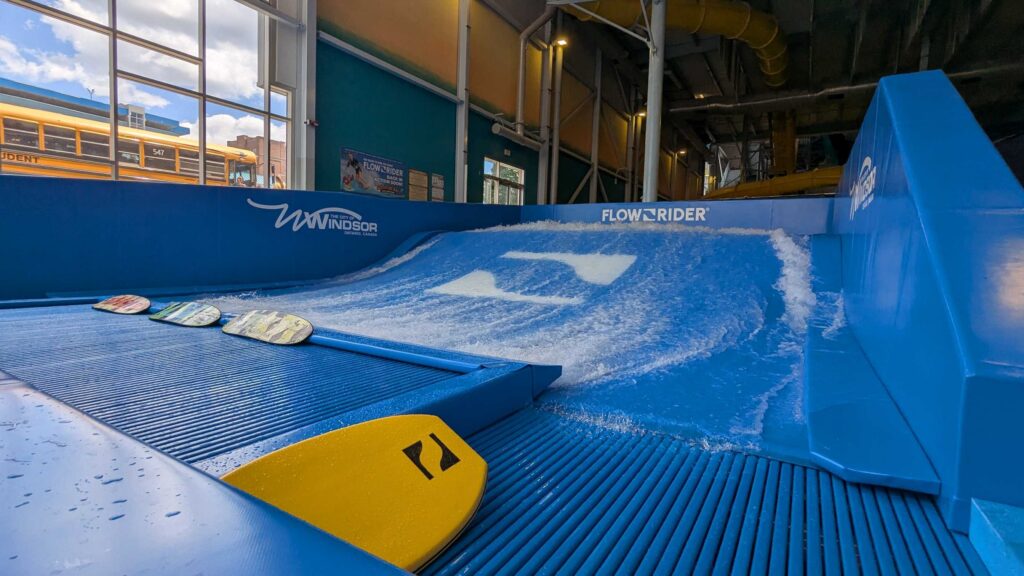 Indoor artificial surfing wave with blue and white water, several bodyboards in the foreground, and a FlowRider sign on the structure; FlowSurf experience enhanced by windows and visible pipes in the background.