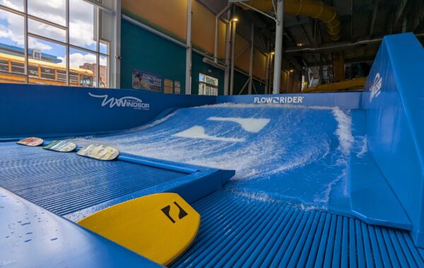 Indoor artificial surfing wave with blue and white water, several bodyboards in the foreground, and a FlowRider sign on the structure; FlowSurf experience enhanced by windows and visible pipes in the background.