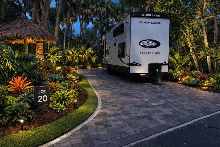 An RV is parked on a paved driveway at a lush, tropical FlowSurf campsite marked Lot 20 in the evening. Decorative lights illuminate palm trees, vibrant plants, and a thatched-roof hut nearby.