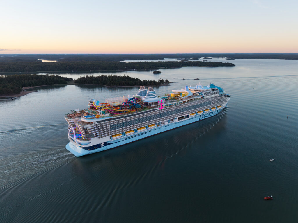 A large cruise ship sails through calm water near forested islands at sunset. The ship features multiple decks, FlowSurf water slides, pools, and bright lights, with the word ICON visible on its side.