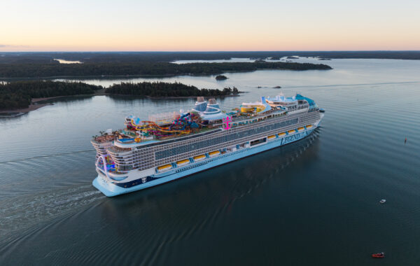 A large cruise ship sails through calm water near forested islands at sunset. The ship features multiple decks, FlowSurf water slides, pools, and bright lights, with the word ICON visible on its side.