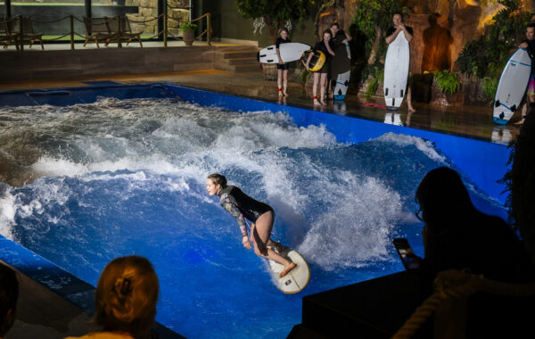 A person surfs on an indoor FlowSurf artificial wave while several people stand on the edge holding surfboards and others watch from behind a rope barrier. The scene is brightly lit with a lively atmosphere.