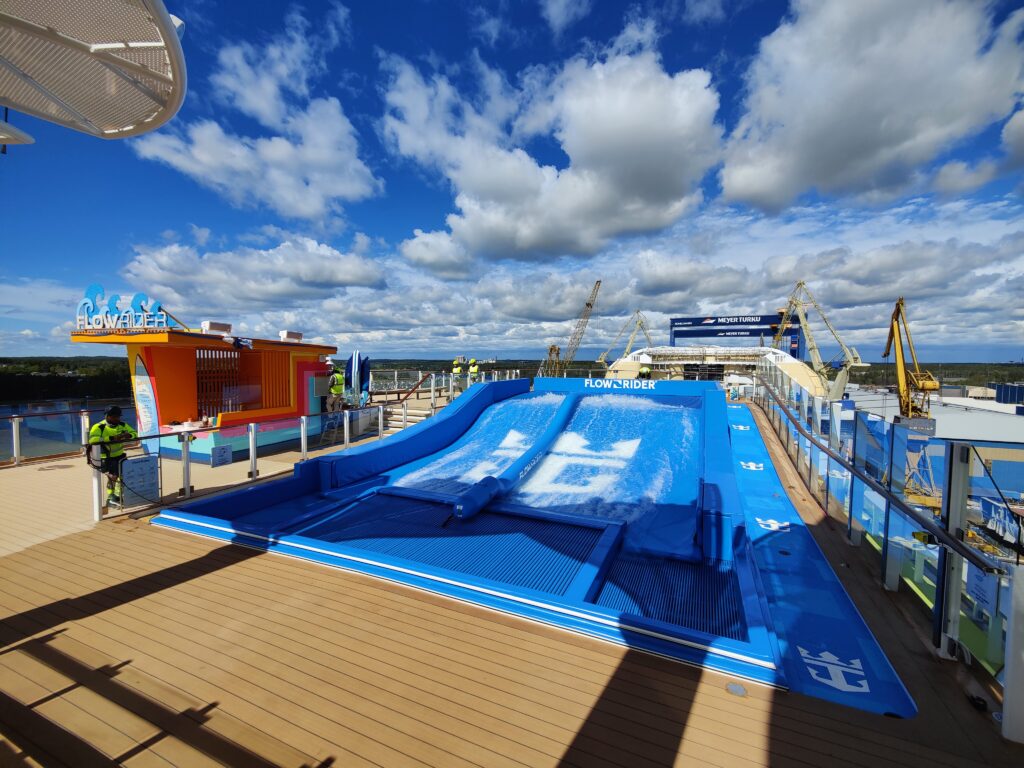 A bright blue FlowSurf surf simulator sits on the deck of a cruise ship under a sunny, partly cloudy sky. Crew members stand nearby as the ship overlooks a body of water and forested shoreline.