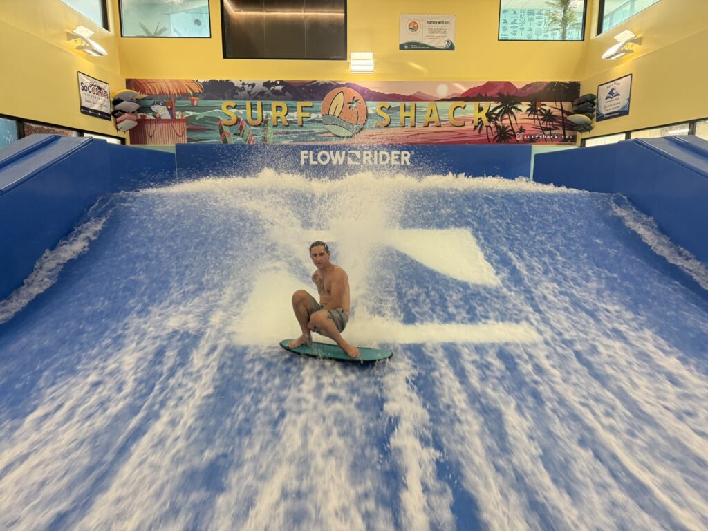 A person rides a wave on an indoor flowrider at Surf Shack, surrounded by bright blue water with surfboards and tropical murals in the background, showcasing skills that could shine at a Flow Tour event.