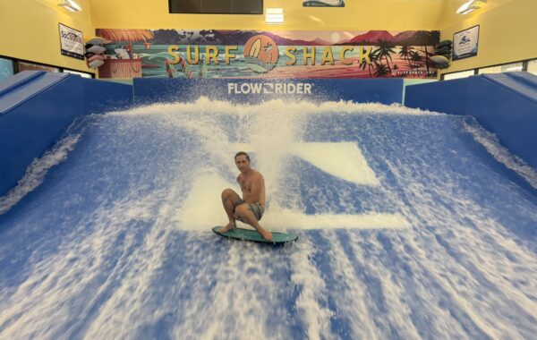 A person rides a wave on an indoor flowrider at Surf Shack, surrounded by bright blue water with surfboards and tropical murals in the background, showcasing skills that could shine at a Flow Tour event.