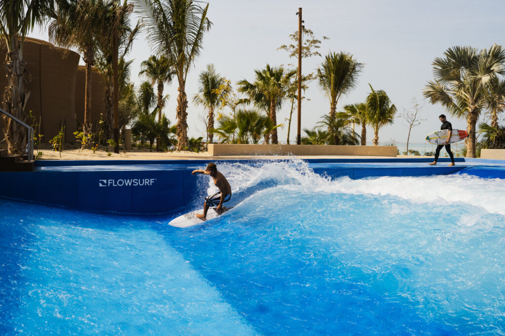 A person surfs an artificial wave at a FlowSurf facility surrounded by palm trees, while another person carrying a surfboard walks in the background.