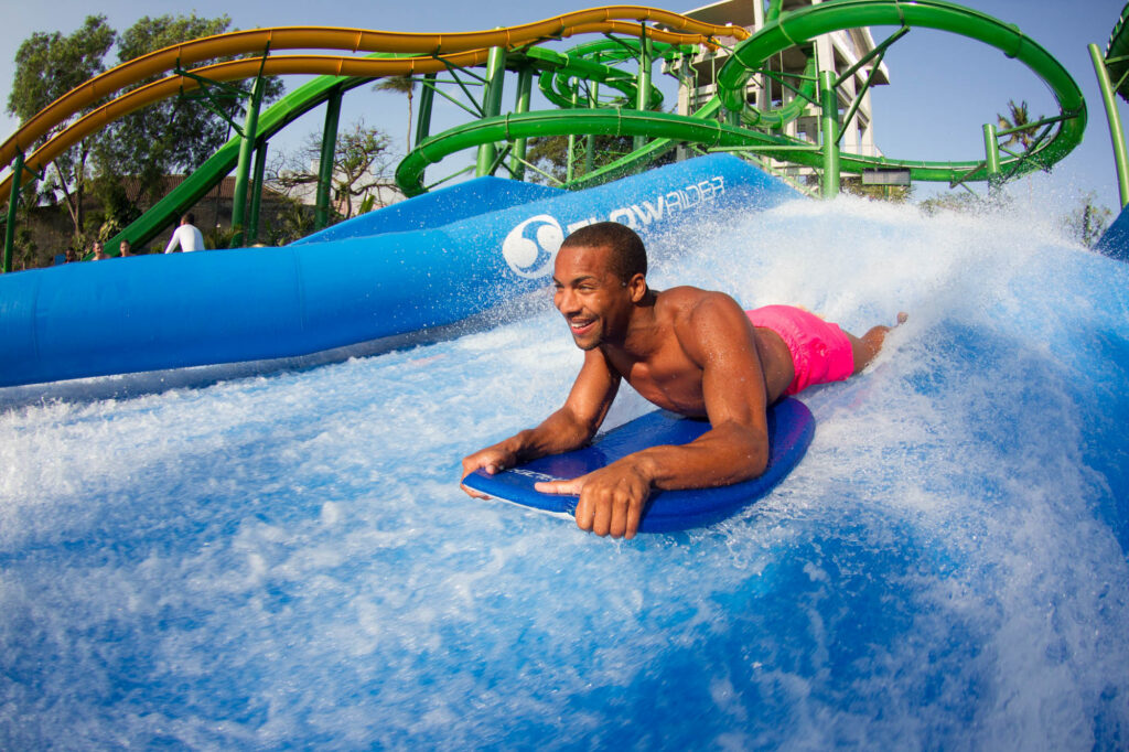 A man in bright pink shorts rides a bodyboard on an artificial wave at Waterbom Bali, smiling as water splashes around him. Green water slides and trees are visible in the background.