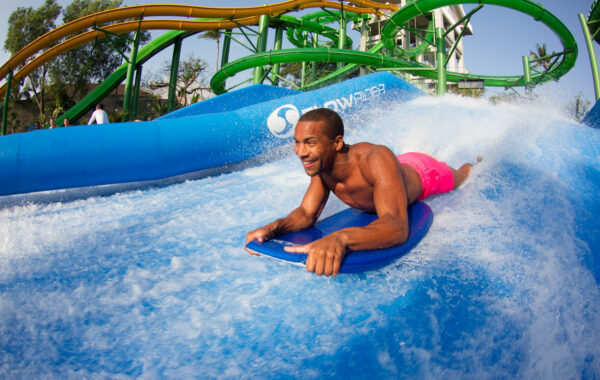 A man in bright pink shorts rides a bodyboard on an artificial wave at Waterbom Bali, smiling as water splashes around him. Green water slides and trees are visible in the background.
