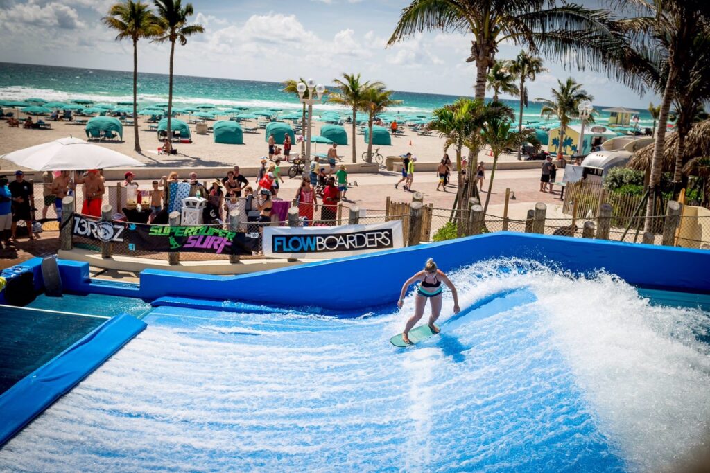 A person rides an artificial wave on a flowboarding machine at Waterbom Bali, a popular beach resort, while people watch from behind a fence. The ocean, palm trees, and beach tents are visible in the background.