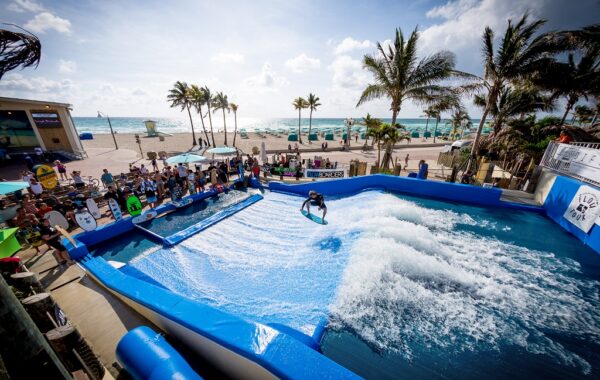 A person surfs on a simulated wave at Waterbom Bali’s outdoor wave pool near the beach, surrounded by spectators, palm trees, and a blue sky with scattered clouds.
