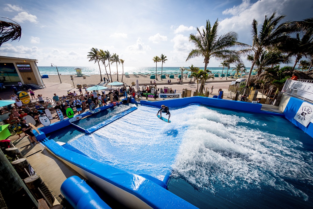 A person surfs on a simulated wave at Waterbom Bali’s outdoor wave pool near the beach, surrounded by spectators, palm trees, and a blue sky with scattered clouds.