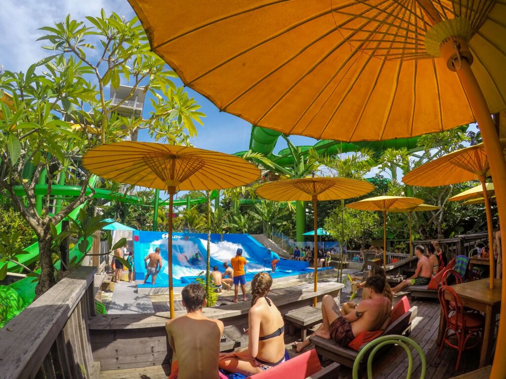 People watch and surf on artificial waves at Waterbom Bali, a tropical water park shaded by large yellow umbrellas, with lush green trees and a relaxed, lively atmosphere.