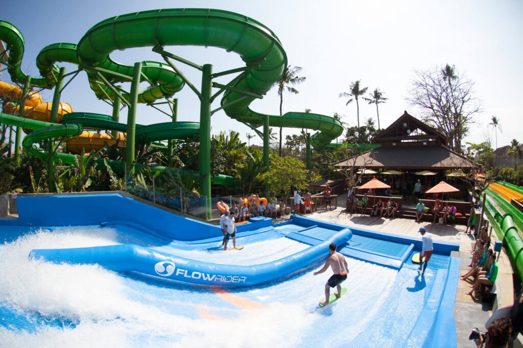 People surf on an artificial wave machine at a waterpark, with winding green waterslides and palm trees in the background at Waterbom Bali. Others watch from shaded seating areas nearby. The scene is bright and lively.