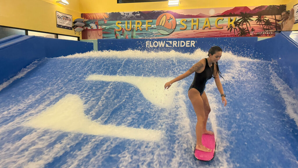 A woman in a black swimsuit rides a pink board on an indoor surfing simulator at Surf Shack during the Flow Tour, with colorful murals and artificial waves in the background.