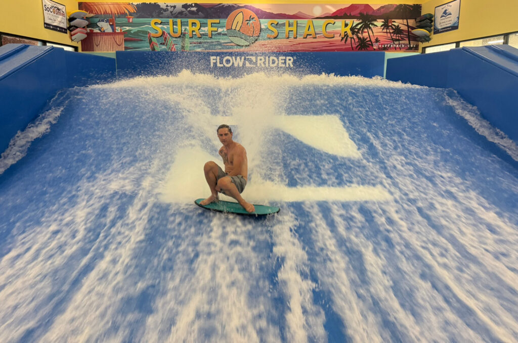 A person in swim trunks rides a surfboard on an indoor wave simulator at Surf Shack, surrounded by blue water and tropical decor.