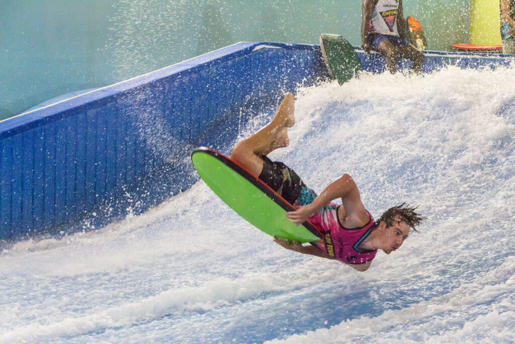 A person in a pink shirt falls off a bright green bodyboard while riding artificial indoor waves during a Flow Tour event, with water splashing around them.