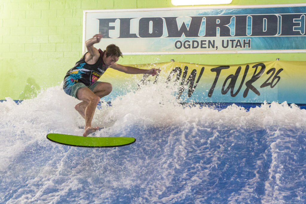 A surfer rides an artificial wave indoors at a FlowRider facility in Ogden, Utah. He balances on a bright green board with water splashing around him, showcasing his skills during the Flow Tour. A Flow Tour 26 sign appears in the background.