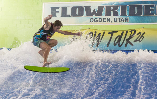 A surfer rides an artificial wave indoors at a FlowRider facility in Ogden, Utah. He balances on a bright green board with water splashing around him, showcasing his skills during the Flow Tour. A Flow Tour 26 sign appears in the background.