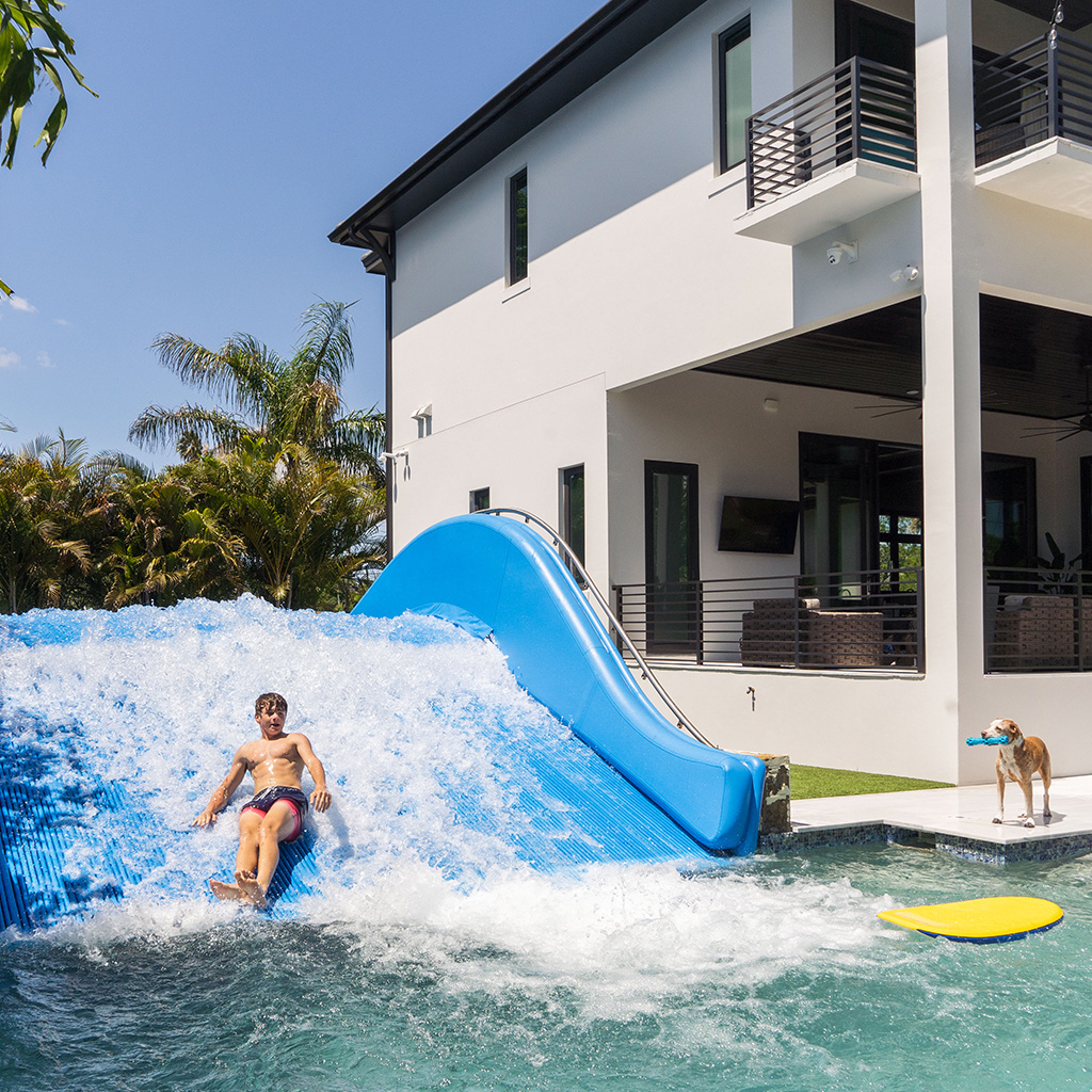 A person rides an artificial wave on a blue wave machine next to a modern white house; a dog stands nearby on the grass next to a yellow surfboard. Palm trees and clear blue sky are in the background.