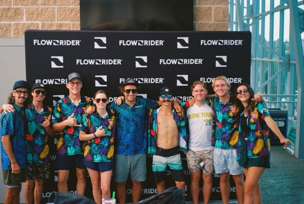 A group of nine people in tropical shirts pose and smile together in front of a FlowRider-branded backdrop, some giving thumbs up. They appear to be at a sporting or surfing event, likely part of the exciting Flow Tour.