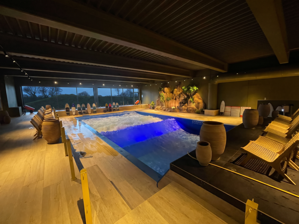 Indoor surf pool with artificial waves, surrounded by lounge chairs and large decorative pots. Tropical plants and surfboards decorate one wall, evoking the signature FlowSurf vibe. Large windows show an outdoor evening view, while warm ambient lighting fills the space.