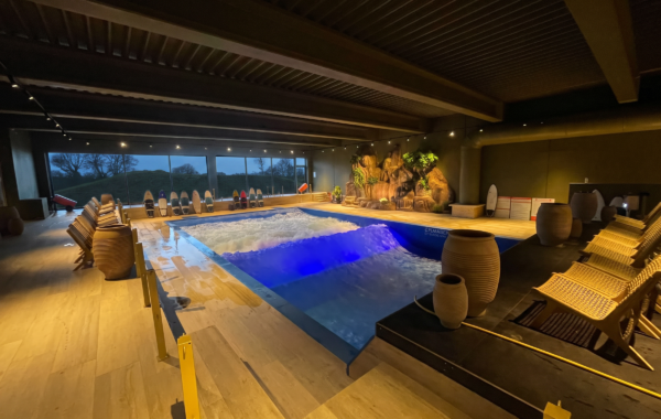 Indoor surf pool with artificial waves, surrounded by lounge chairs and large decorative pots. Tropical plants and surfboards decorate one wall, evoking the signature FlowSurf vibe. Large windows show an outdoor evening view, while warm ambient lighting fills the space.