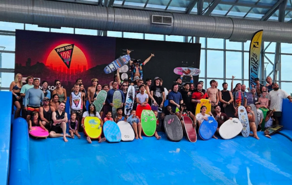 A large group of people posing with bodyboards and surfboards on an indoor wave pool during the Flow Tour. Some are standing, others are sitting, and a few hold their boards above their heads, smiling for the camera.