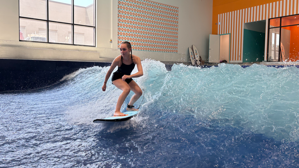 A person in a black swimsuit surfs an indoor wave pool, balancing on a blue surfboard during the Flow Tour. There are surfboards and colorful wall patterns in the background.