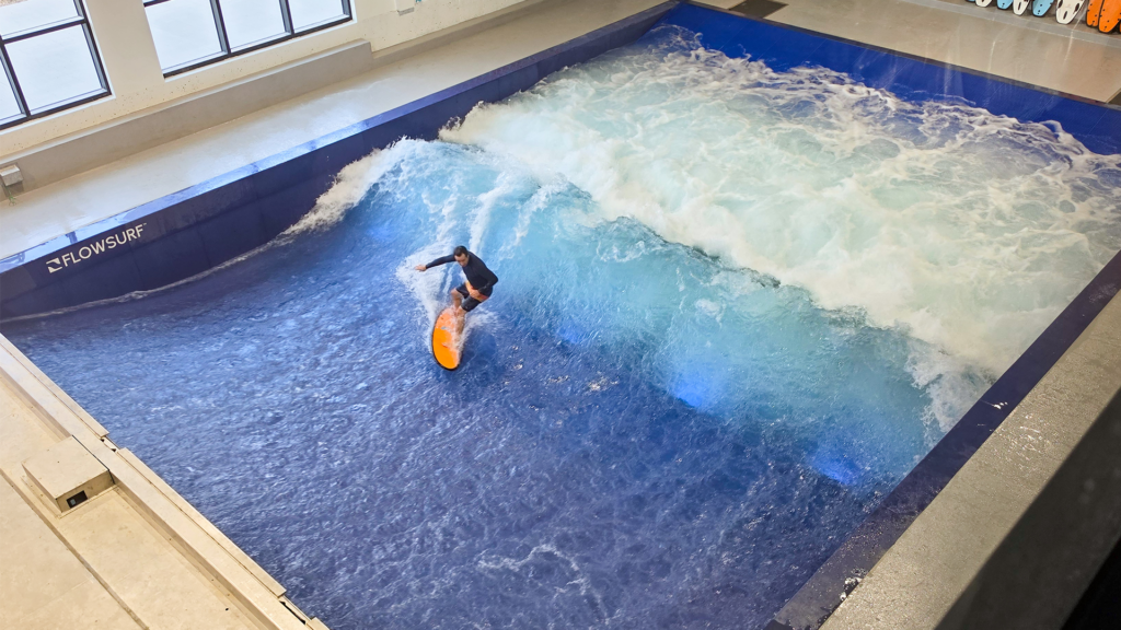 A person surfs on an artificial indoor wave at a Flow Tour facility with blue water and white foam, surrounded by walls and windows.