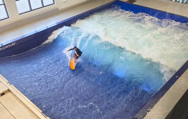 A person surfs on an artificial indoor wave at a Flow Tour facility with blue water and white foam, surrounded by walls and windows.