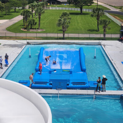 A wave simulator in a pool with people surfing on the FlowRider Double, some watching from the edge, and an instructor assisting. Green grass, trees, and a road are visible in the background.
