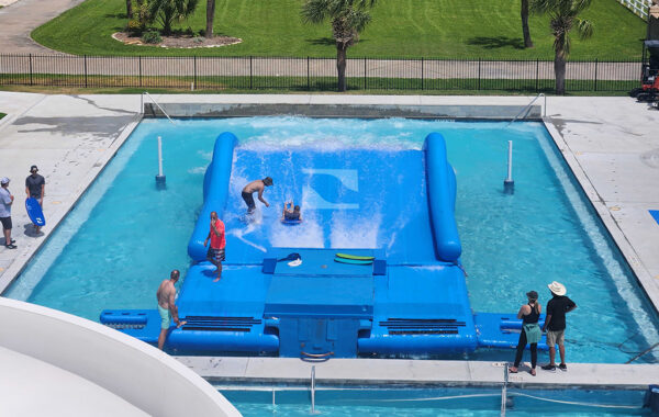 A wave simulator in a pool with people surfing on the FlowRider Double, some watching from the edge, and an instructor assisting. Green grass, trees, and a road are visible in the background.