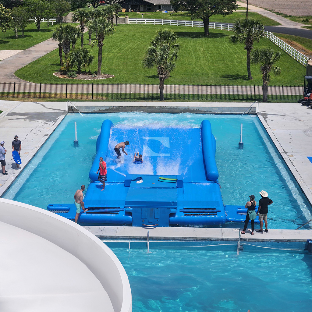 A wave simulator in a pool with people surfing on the FlowRider Double, some watching from the edge, and an instructor assisting. Green grass, trees, and a road are visible in the background.