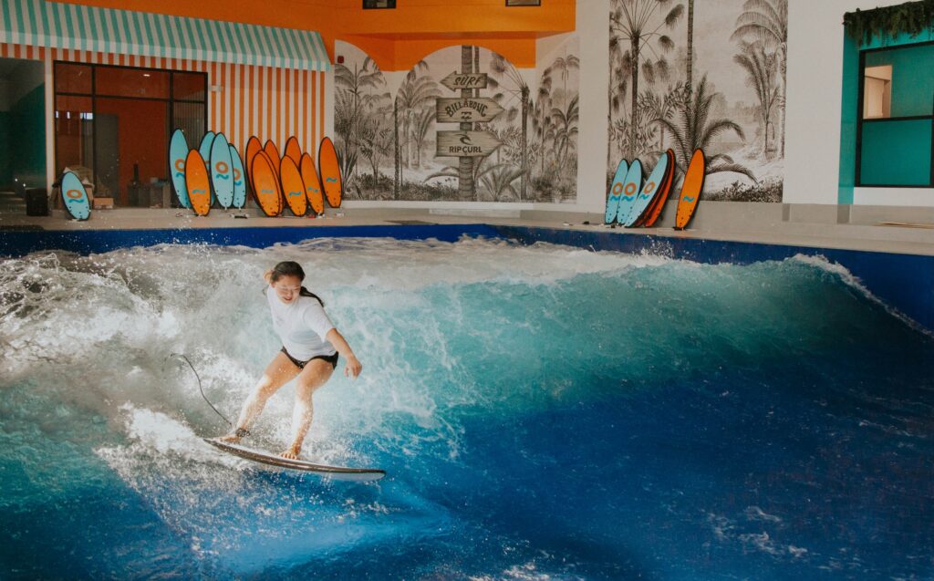 A person surfs on an indoor FlowSurf artificial wave, surrounded by water and several colorful surfboards lined up in the background.