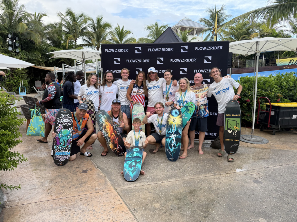 A group of people posing with flowboards in front of a FLOWRIDER backdrop at an outdoor Flow Tour event, surrounded by palm trees and tropical plants.