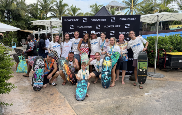 A group of people posing with flowboards in front of a FLOWRIDER backdrop at an outdoor Flow Tour event, surrounded by palm trees and tropical plants.