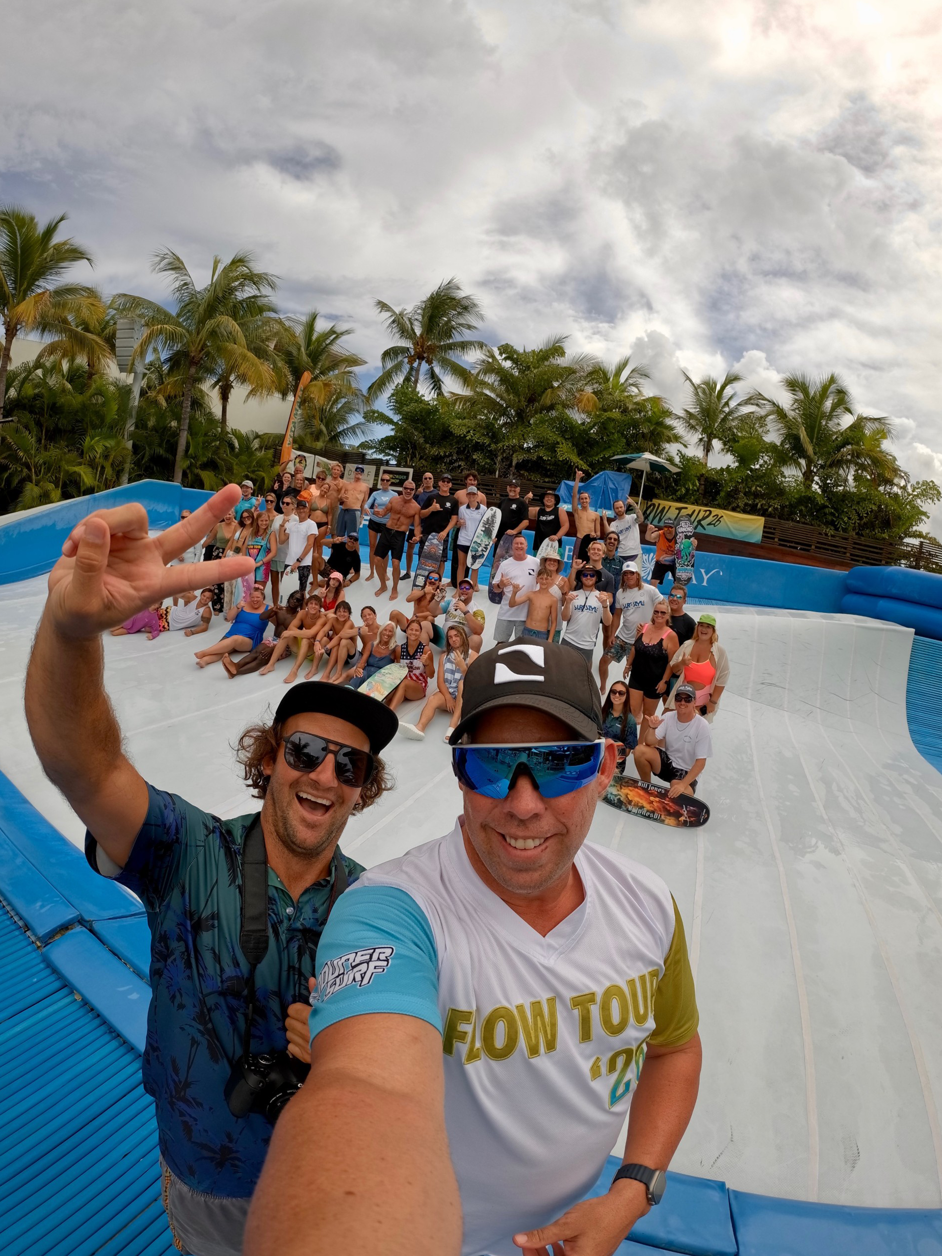 Two men take a selfie in front of a large group posing on an empty wave pool during the Flow Tour, surrounded by palm trees and cloudy skies. Most people are smiling, wearing casual summer clothing, and appear to be at an outdoor event.
