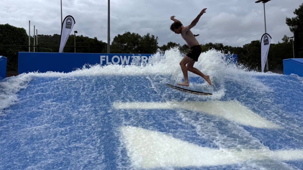 A person surfing on an artificial wave at a FlowRider facility, balancing on a board as water splashes around them under a cloudy sky.