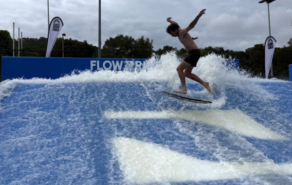 A person surfing on an artificial wave at a FlowRider facility, balancing on a board as water splashes around them under a cloudy sky.